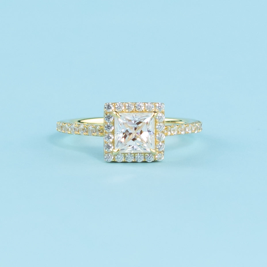 This ring showcases a brilliant princess cut moissanite with a timeless halo setting in warm yellow gold.