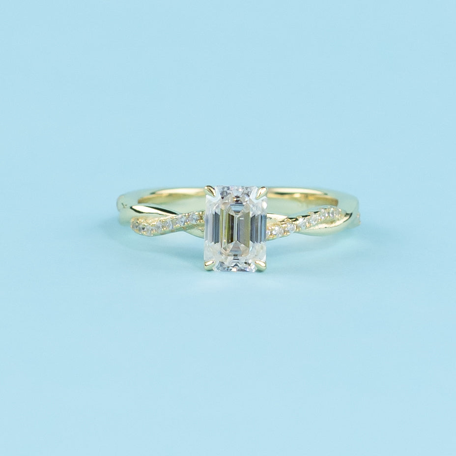 Showcases an elegant emerald cut moissanite set within a flowing infinity design in warm yellow gold.