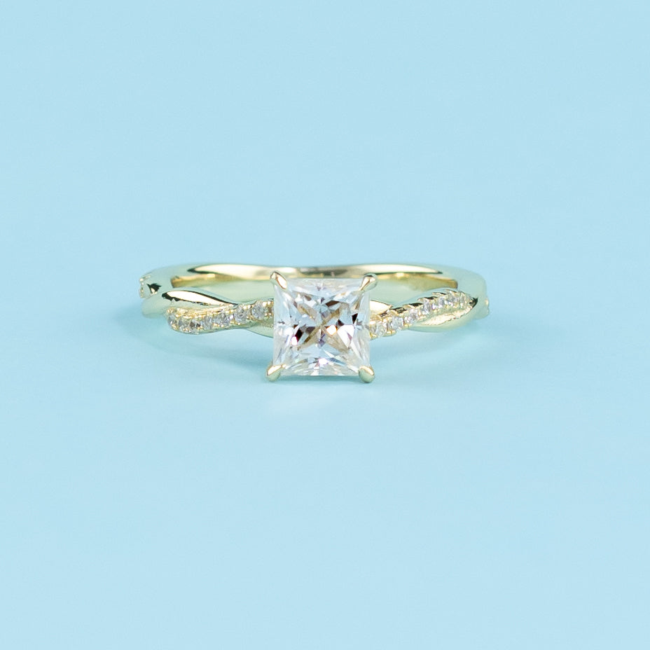 Showcases a beautiful princess cut moissanite set in a warm yellow gold band with a symbolic infinity twist.