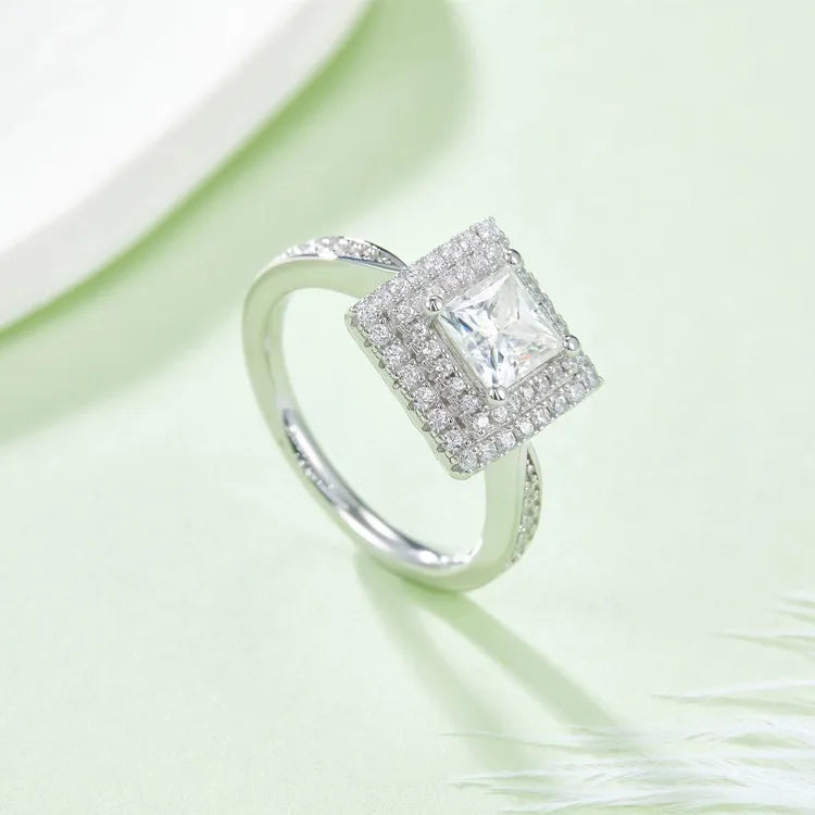 This ring features a 1.00ct princess cut moissanite surrounded by two layers of shimmering stones, creating a radiant look.