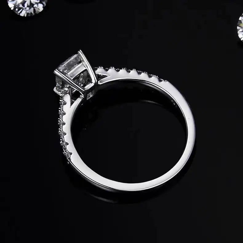 This ring features a brilliant 1.00ct emerald cut moissanite displayed in a traditional basket setting, emphasizing its clean lines.
