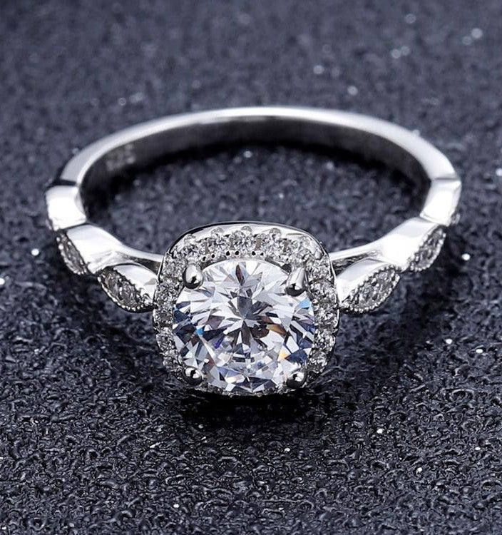 This ring showcases a brilliant 1.00ct moissanite with a timeless halo design, crafted in intricate sterling silver with vintage details.