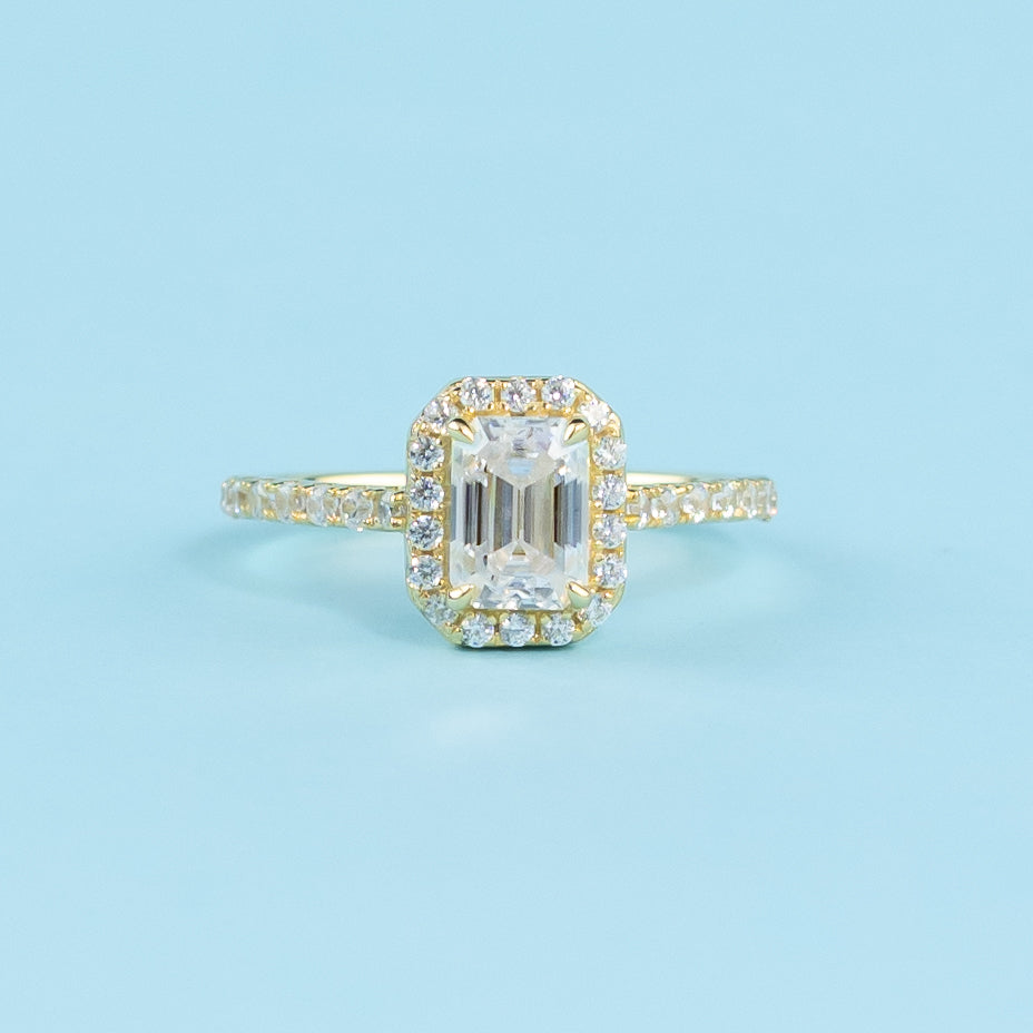 This ring showcases a brilliant emerald cut moissanite with a timeless halo setting in warm yellow gold.