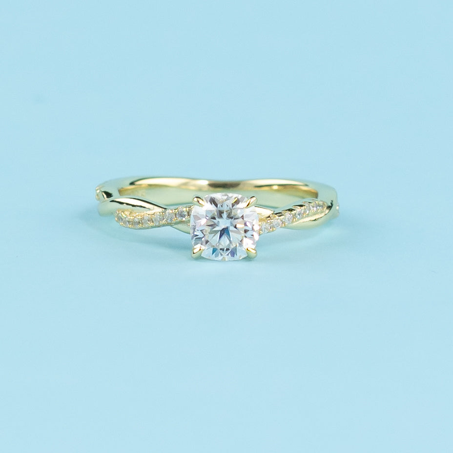 Showcases a beautiful cushion cut moissanite set in a warm yellow gold band with a symbolic infinity twist.